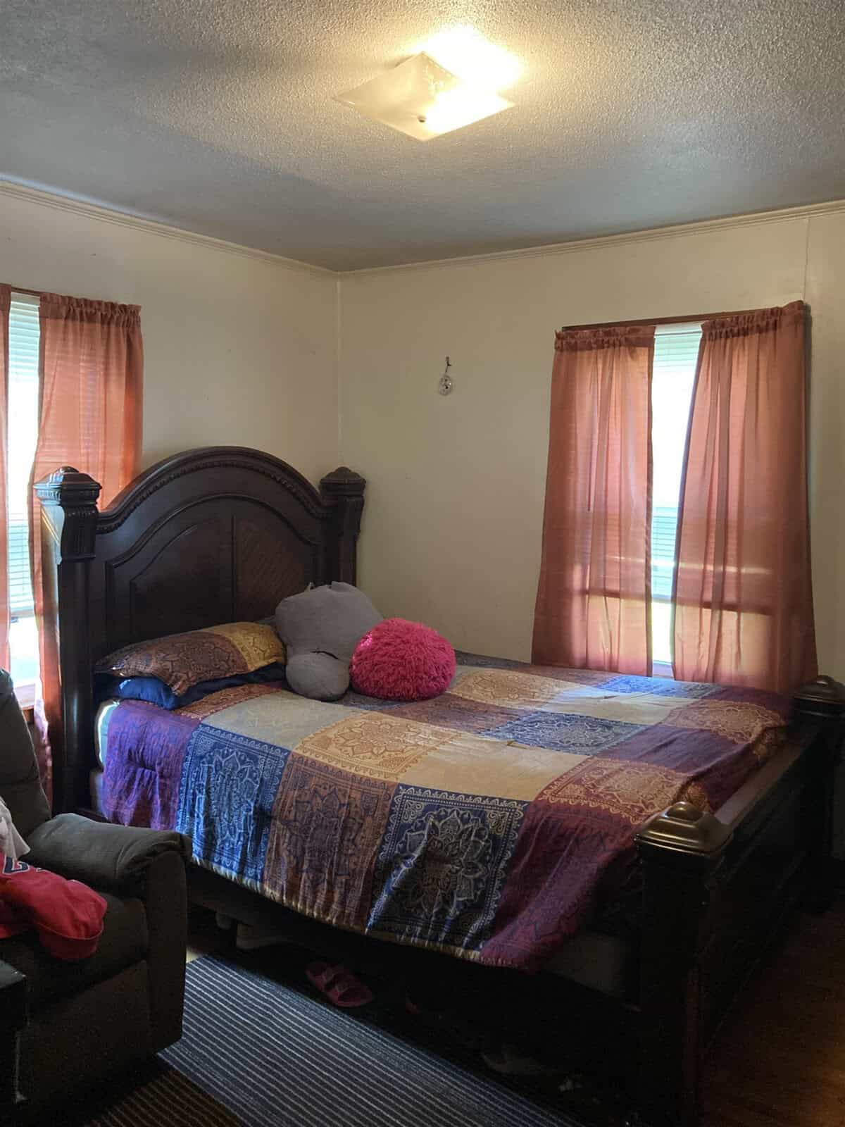 A bedroom with a large bed with a quilted cover, a pink round pillow, and sunlight filtering through sheer curtains.