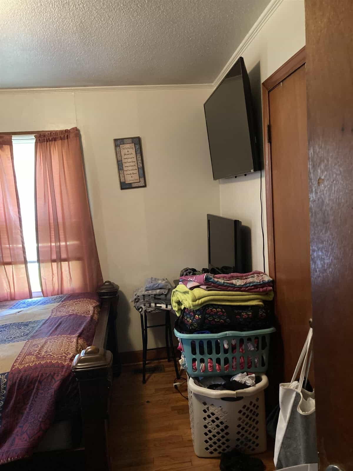 A bedroom with a mounted television, a laundry basket filled with folded clothes, a bed with a blue cover, and orange curtains.