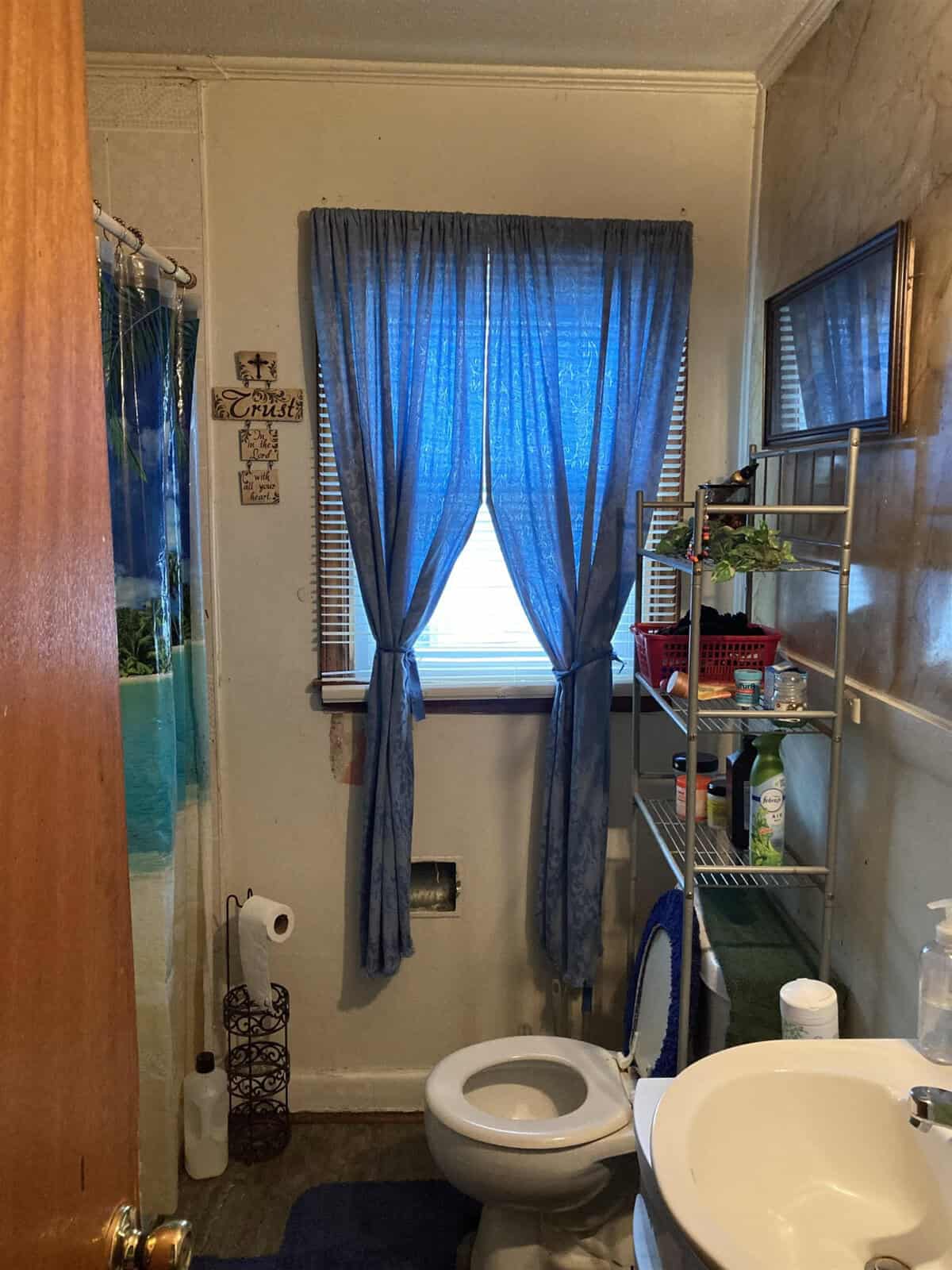 A small bathroom with a window dressed in blue curtains, a toilet, sink, and various toiletries and bathroom supplies.
