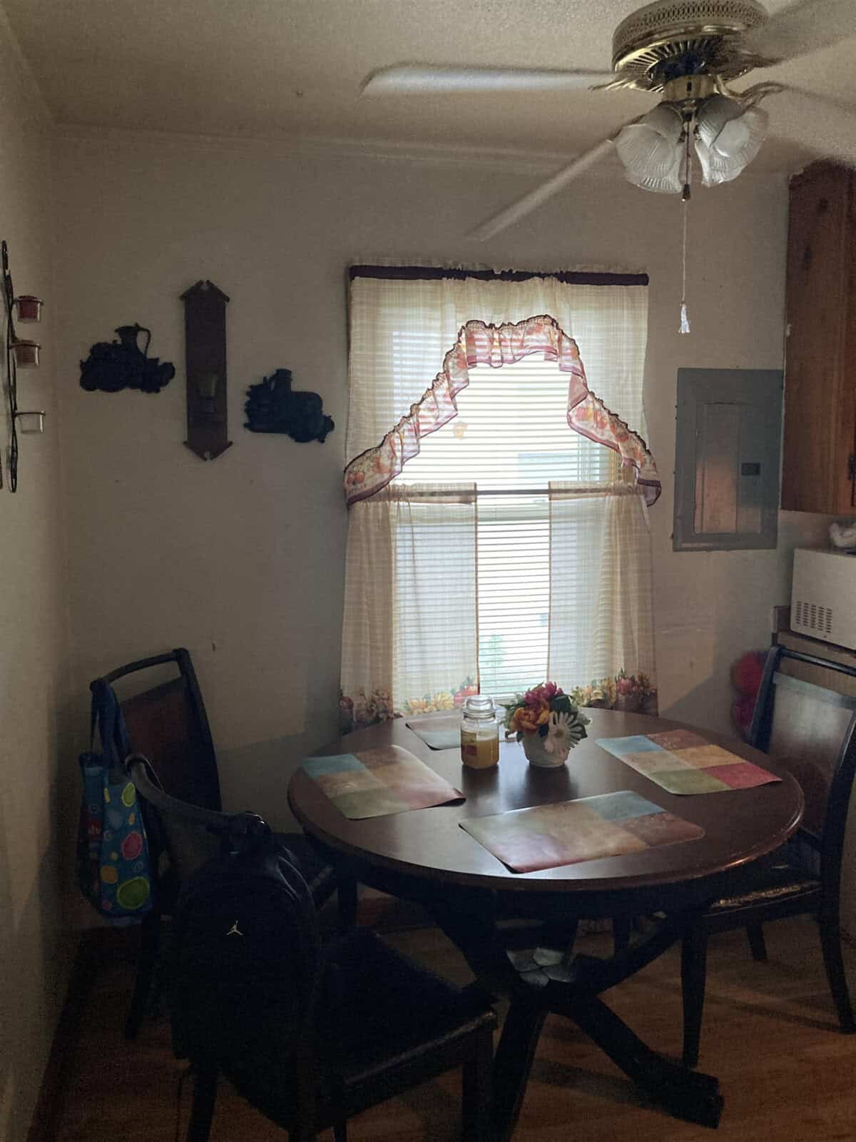 A dimly lit dining room with a round wooden table set with placemats, a ceiling fan above, and a window dressed with ruffled curtains. there are decorative items on the window sill and a backpack hanging on one of the dining chairs.