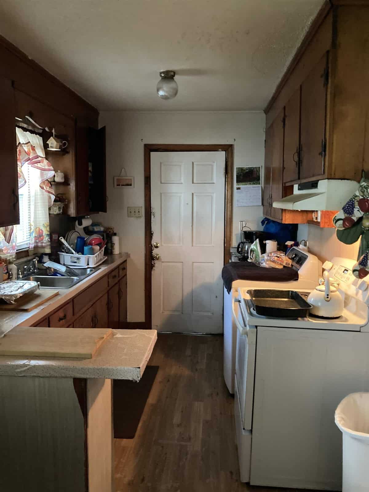 A modest kitchen with wooden cabinetry and a mix of appliances and utensils on the countertops. the room is lit by a single ceiling light, and there's a white door at the far end.