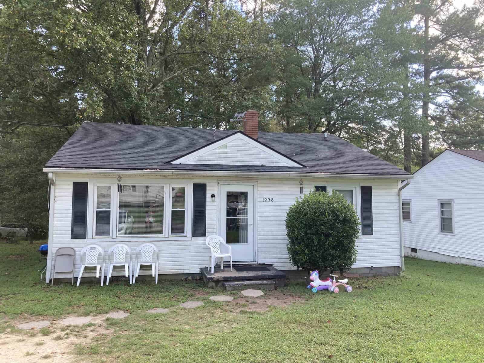 A single-story, white suburban house with a front porch flanked by two plastic chairs and a toy lying on the lawn.