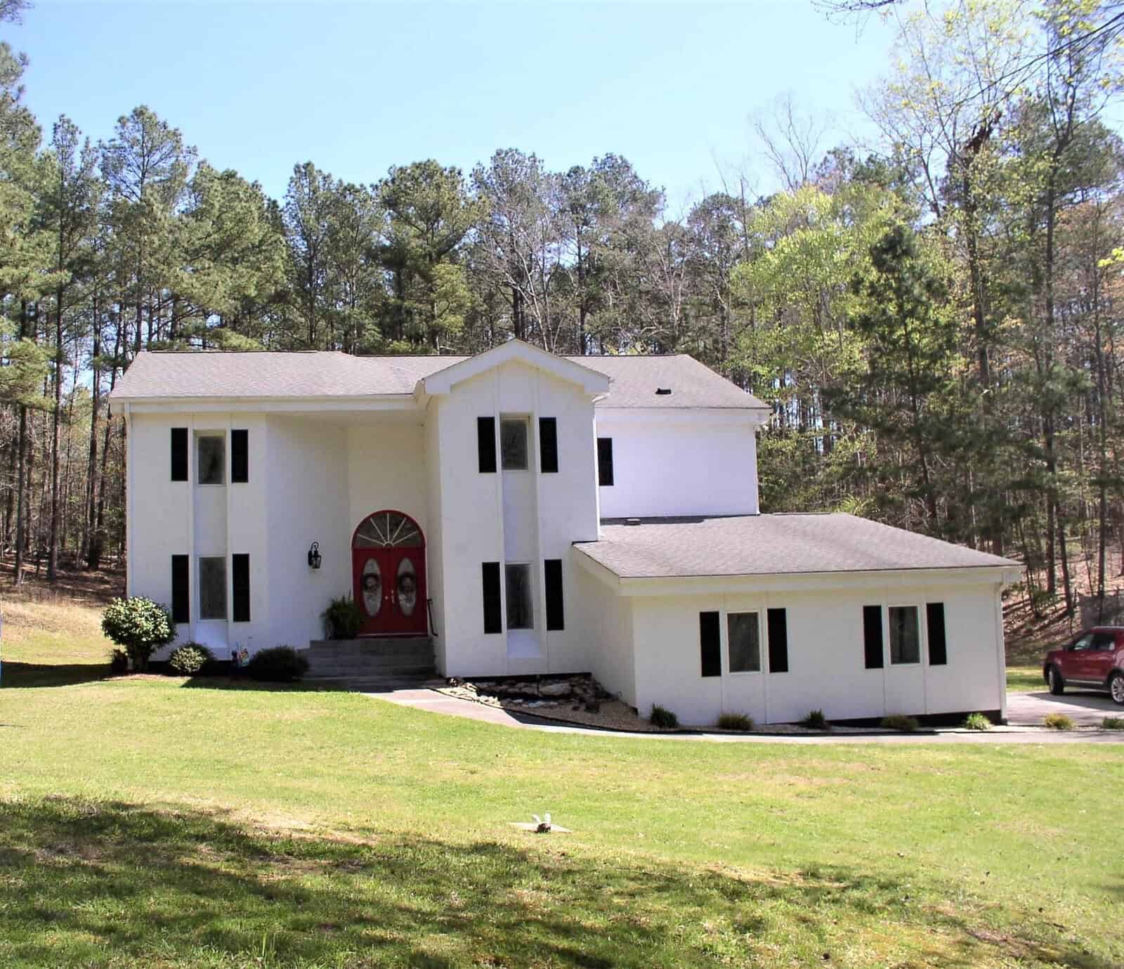 A two-story white house with a red front door, an attached garage, and a car parked in the driveway, set against a backdrop of trees under a clear sky.