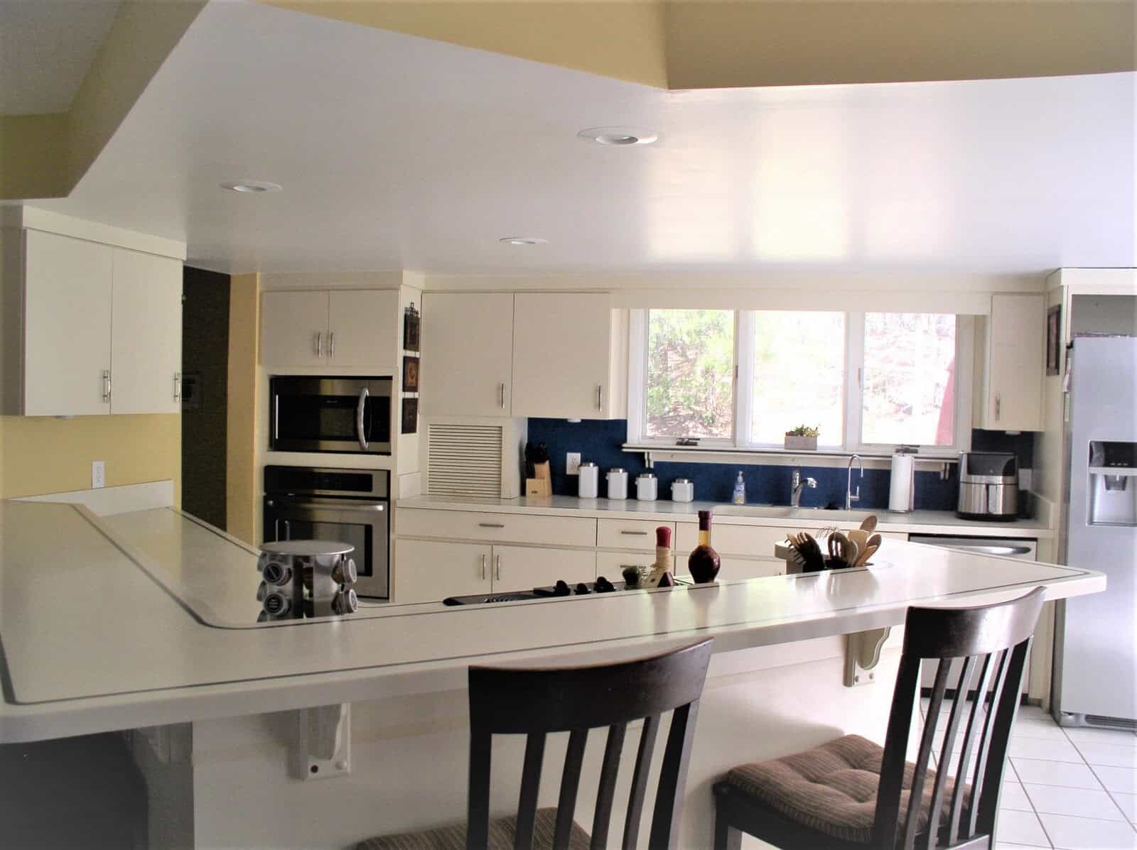 A bright, modern kitchen with white countertops, bar seating, stainless steel appliances, and blue accents visible through a pass-through window.