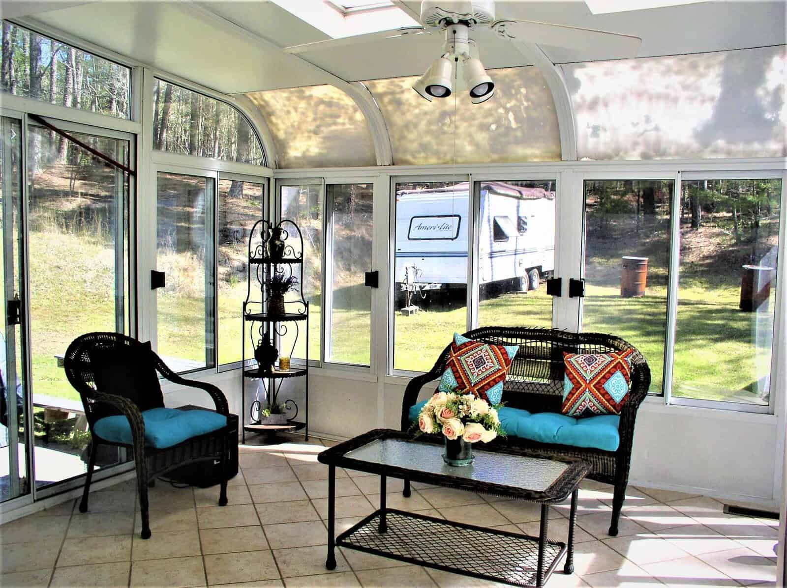 Sunlit enclosed porch with a seating area, featuring a black sofa and two chairs with turquoise cushions, a coffee table, and a plant stand, overlooking a yard with a visible camper trailer through the windows.
