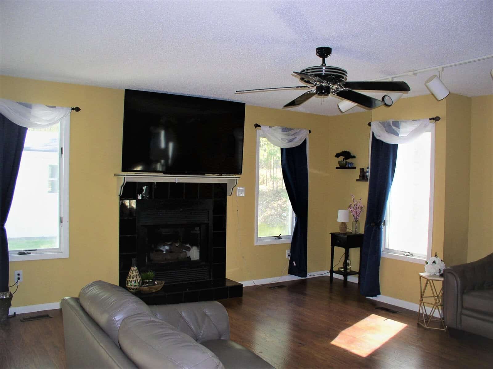 A well-lit living room with yellow walls, a mounted television above a black fireplace, a beige sofa, hardwood floors, and windows dressed with white and blue curtains.