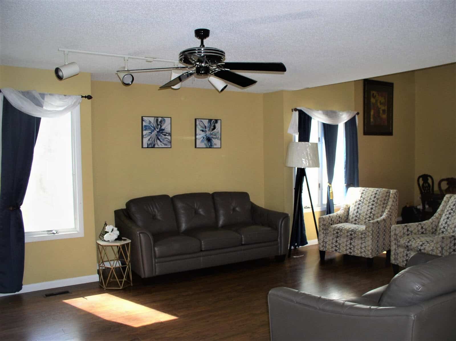 A well-lit living room featuring a dark leather sofa, two patterned armchairs, a ceiling fan, a floor lamp, and wall art, with natural light coming in from a large window adorned with curtains.