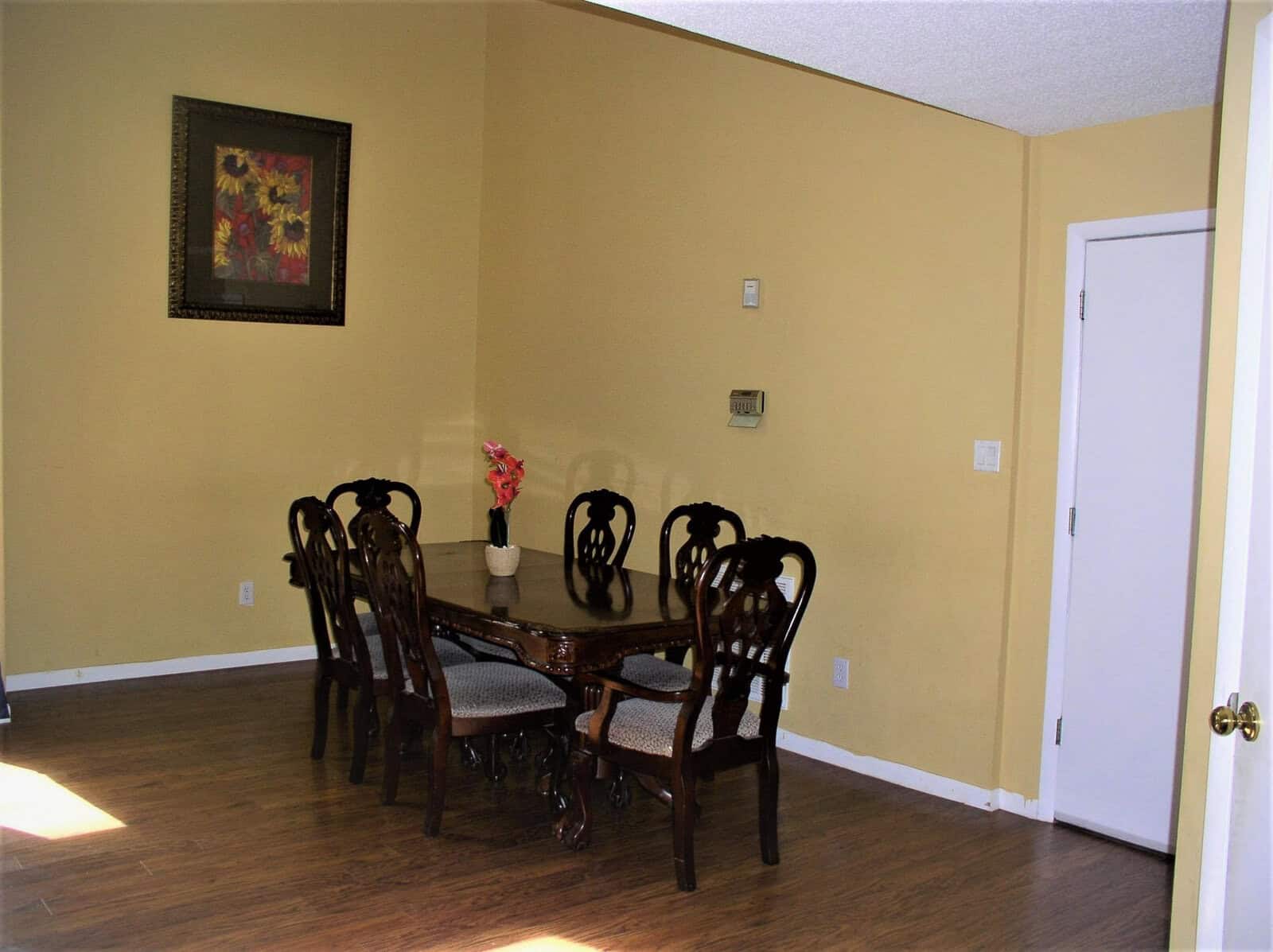 A sparsely furnished dining room with a wooden table and six chairs, beige walls, a small framed artwork, and a vase of flowers on the table.