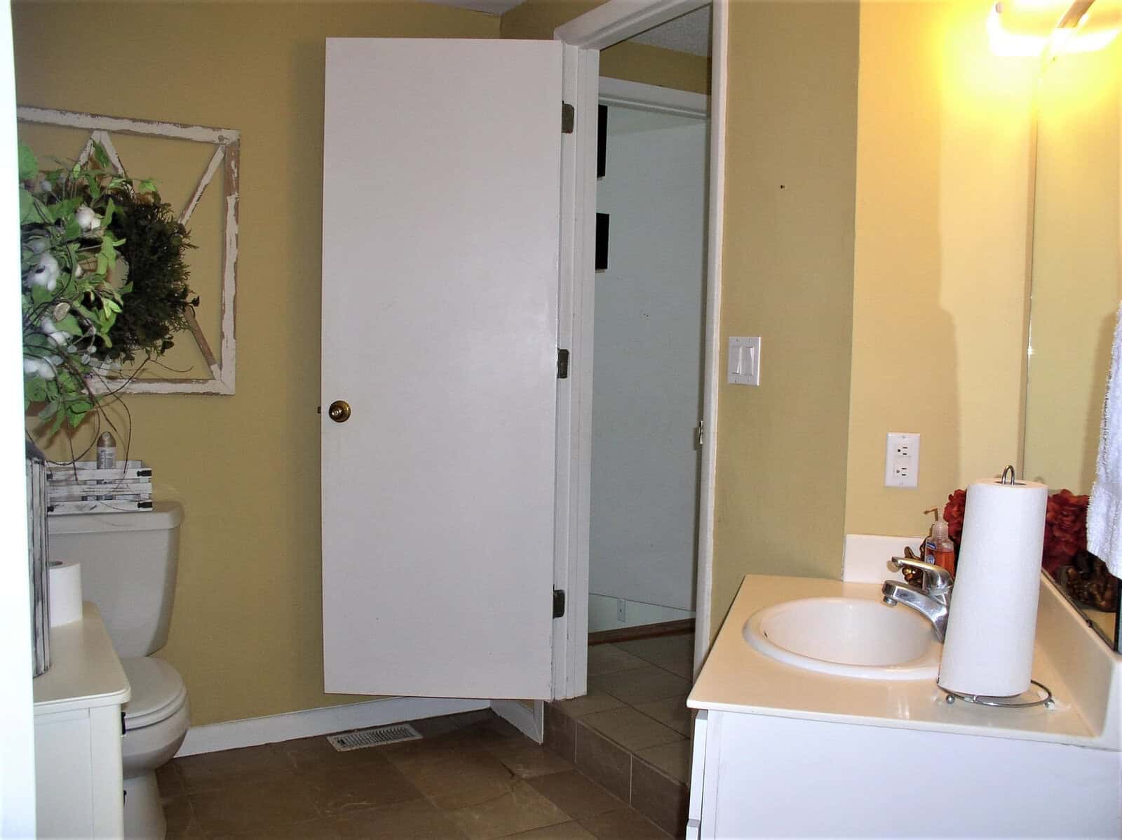 A well-lit bathroom with an open door, featuring a toilet on the left, a sink with a mirror on the right, and tiled flooring. decorative elements include a hanging plant and red accent pieces on the sink countertop.