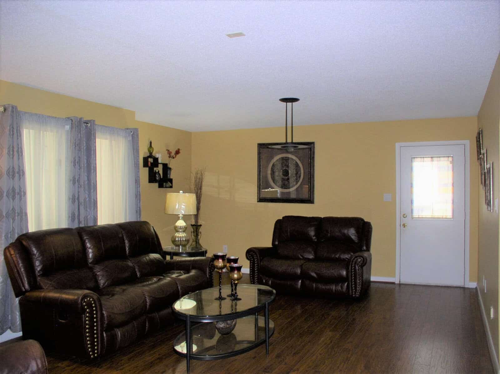 A well-lit living room featuring a brown leather sofa set, a glass-top coffee table, and light-colored walls with decorative items and curtains.