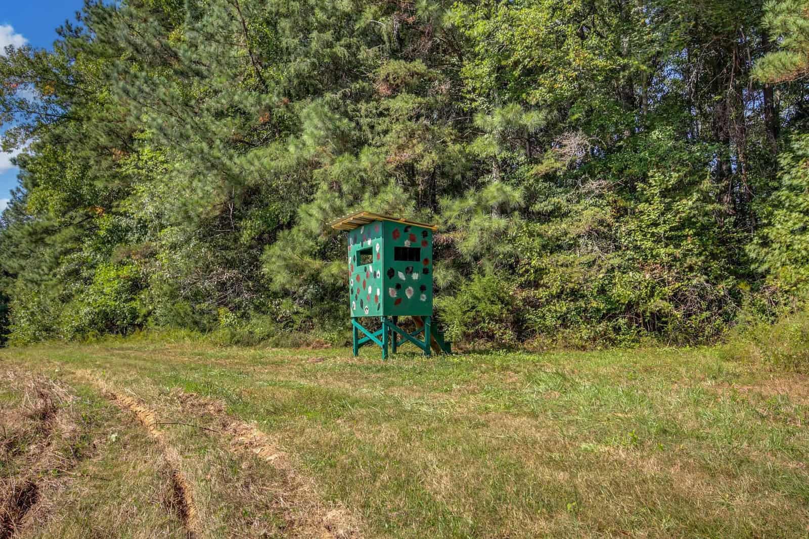 A green hunting blind with camouflage patterns stands on a grassy field in front of a dense forest. The structure is elevated, with a small roof, surrounded by trees and under a blue sky.