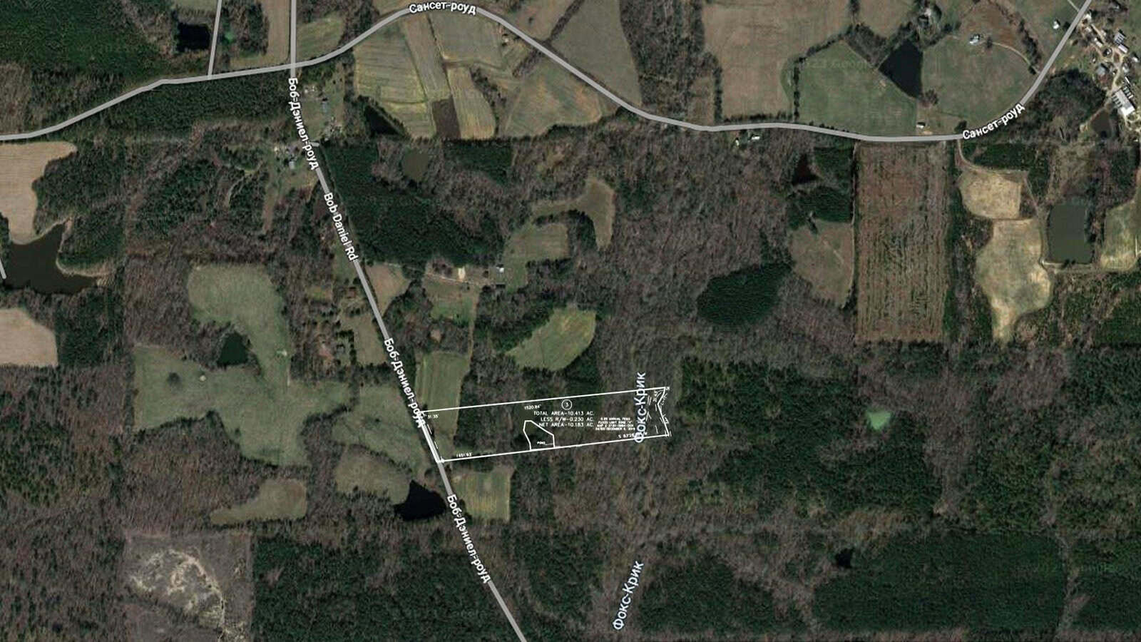 Aerial view of a rural area with forests and farmland. A marked plot of land is visible, showing divisions and measurements. Roads border the area, intersecting at various points. The landscape includes patches of open fields and dense woodland.
