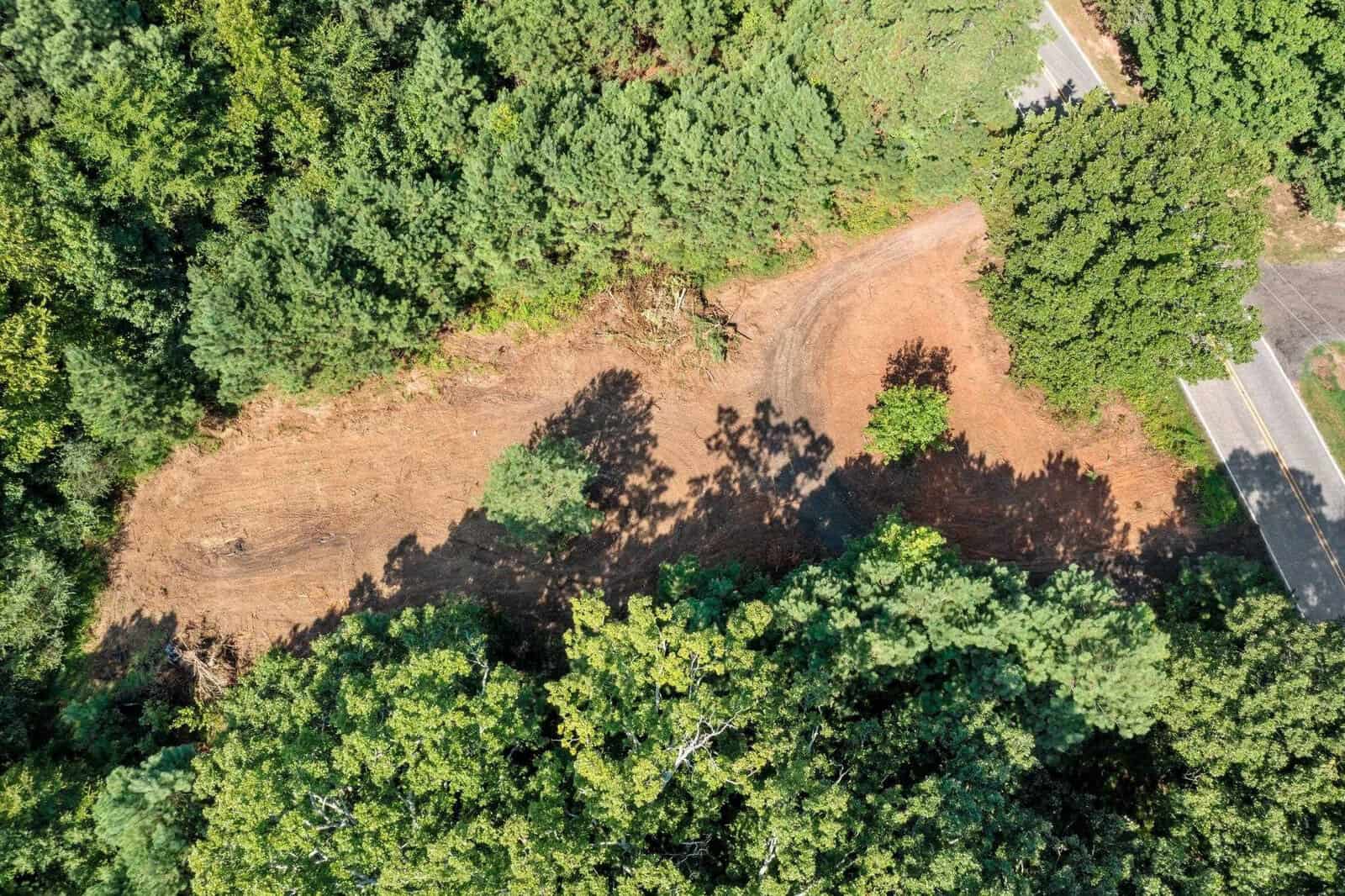 Aerial view of a cleared dirt area surrounded by dense green trees. A narrow road runs along the right side, partially visible. The shadows of the trees fall over the cleared section, creating contrasting patterns.