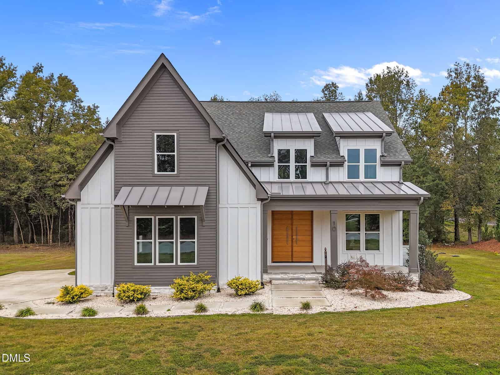 Modern two-story house at 10 Carnation Road in Youngsville, featuring a gray and white exterior, large front windows, double wooden front doors, metal roof, and a landscaped yard with shrubs and a curved walkway. Trees and blue sky complete the view.