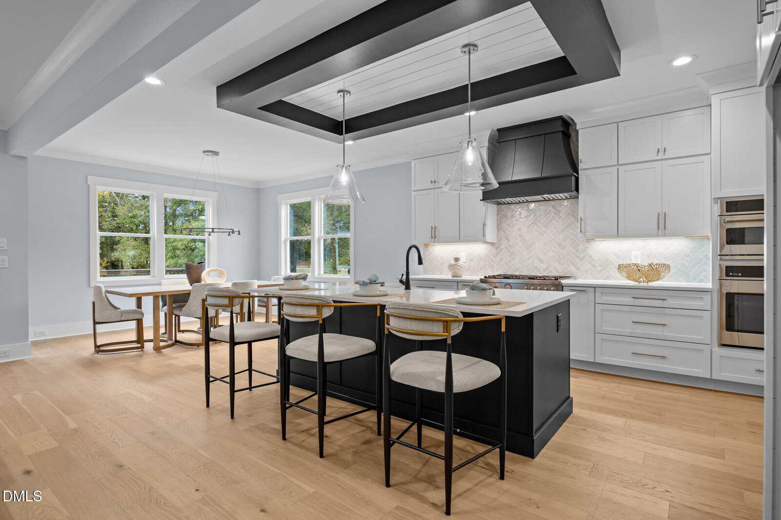 Modern kitchen with a large black island, three bar stools, white cabinets, stainless steel appliances, pendant lights, and a dining area with windows overlooking greenery. Light wood flooring throughout the space.