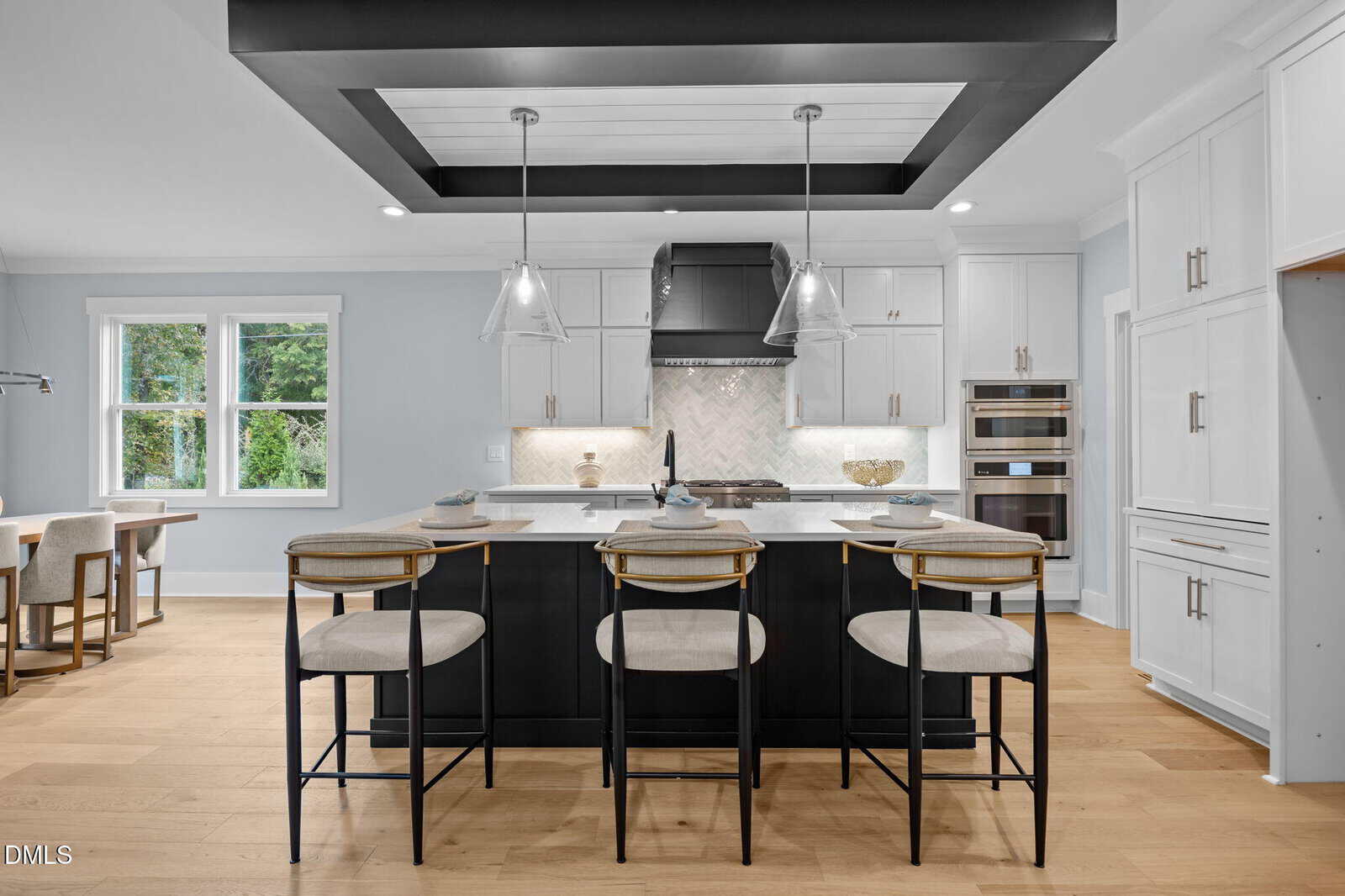 Modern kitchen with a large black island, three barstools, pendant lights, white cabinets, stainless steel appliances, a chevron tile backsplash, and natural light from a window on the left.