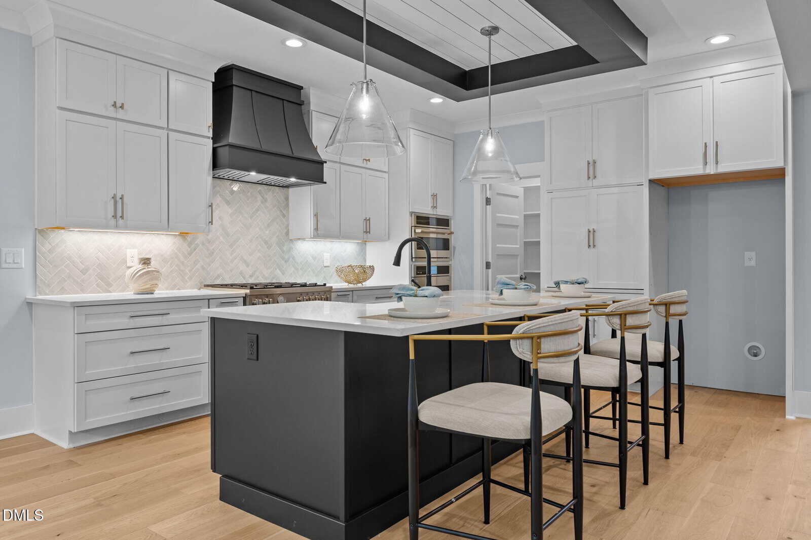 Modern kitchen with white cabinets, black island, pendant lights, and three barstools with light upholstery. Features built-in oven, gas stove, open shelving, and light wood flooring for a clean, contemporary look.
