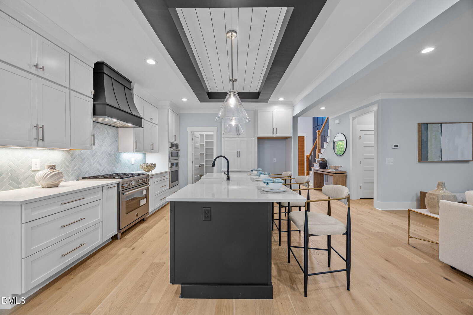 Modern kitchen with white cabinets, stainless steel appliances, black range hood, large island with sink and barstools, light wood flooring, pendant lights, and open view into a hallway and living area.