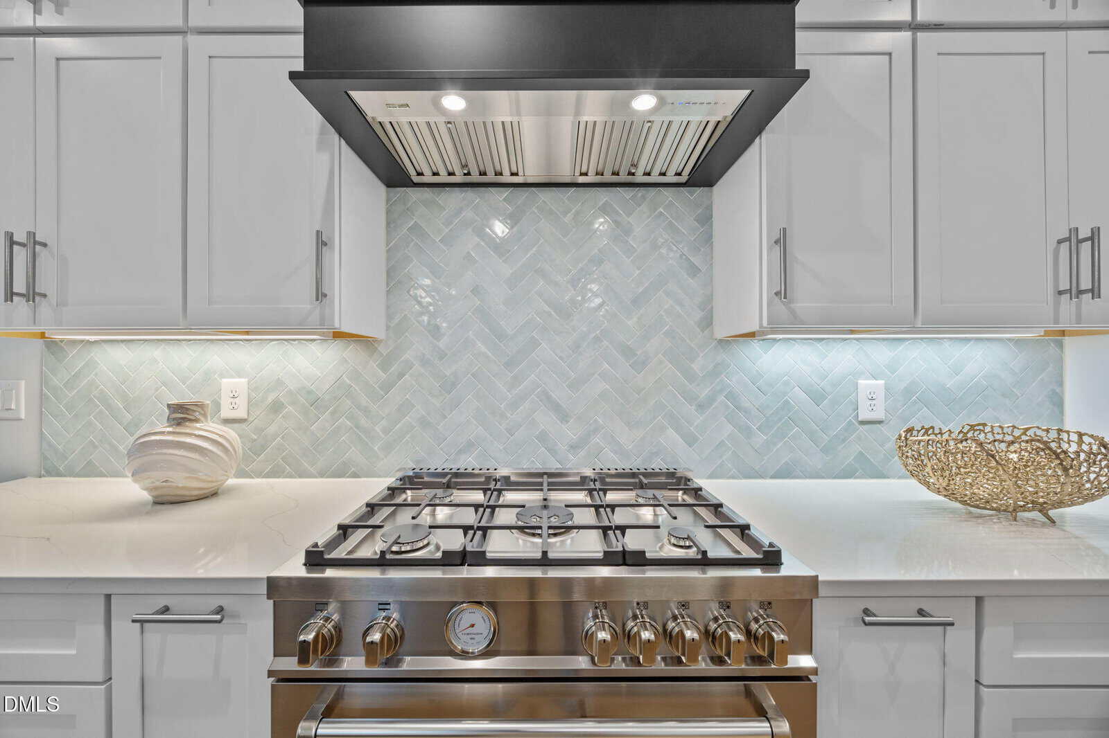 A modern kitchen with a stainless steel gas stove beneath a range hood. The backsplash features a light blue herringbone tile pattern, and decorative items are placed on the white countertops.