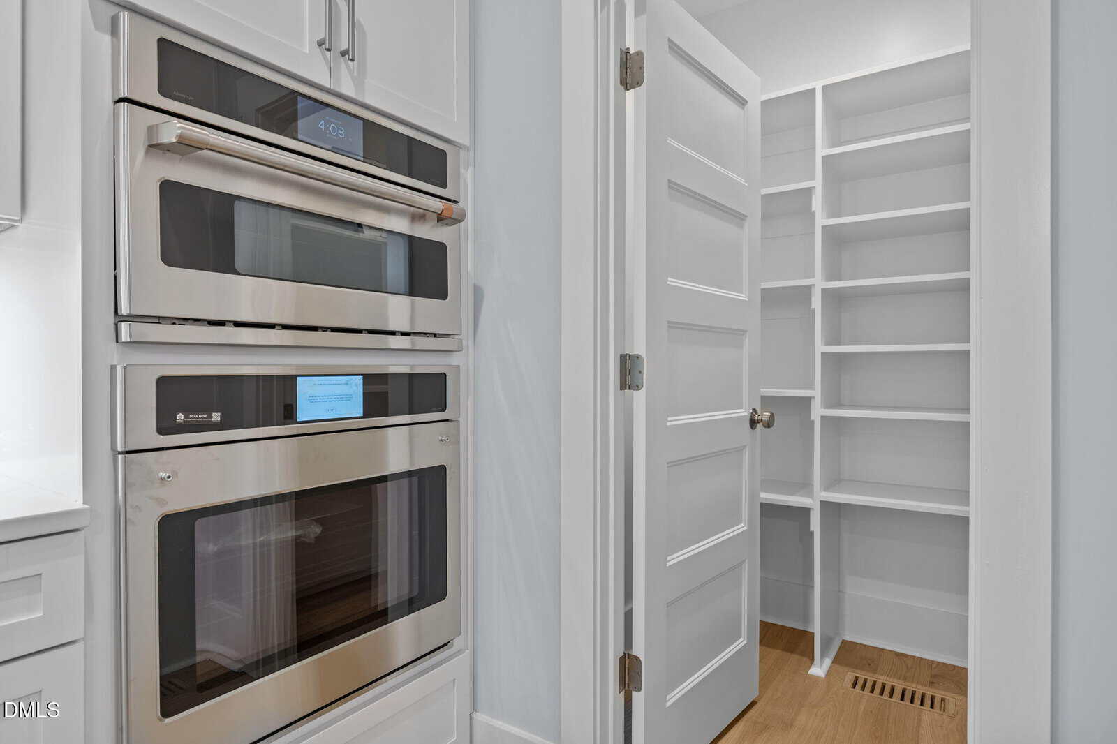 Modern kitchen with built-in stainless steel double ovens on the left and a walk-in pantry with white shelves and a white door on the right, featuring light wood flooring.