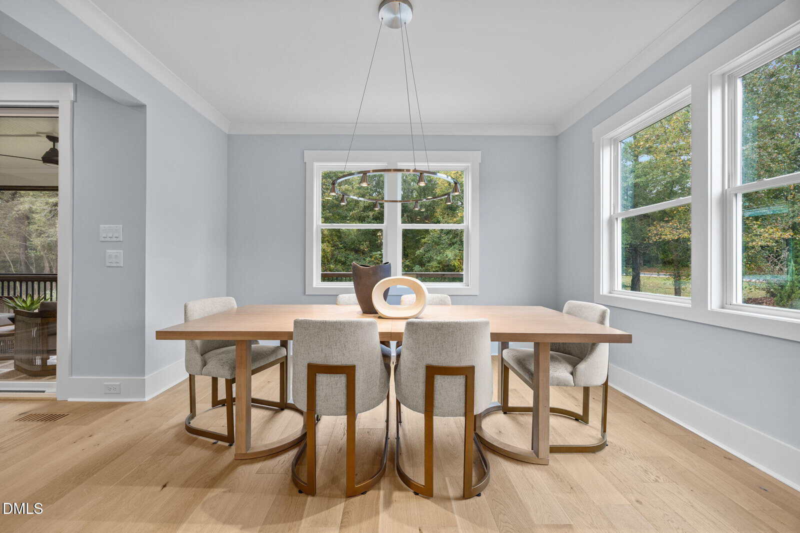 A modern dining room with a wooden table, six upholstered chairs, a round chandelier, and decorative vases. Large windows let in natural light and offer a view of greenery outside. Light wood floors and pale gray walls complete the space.