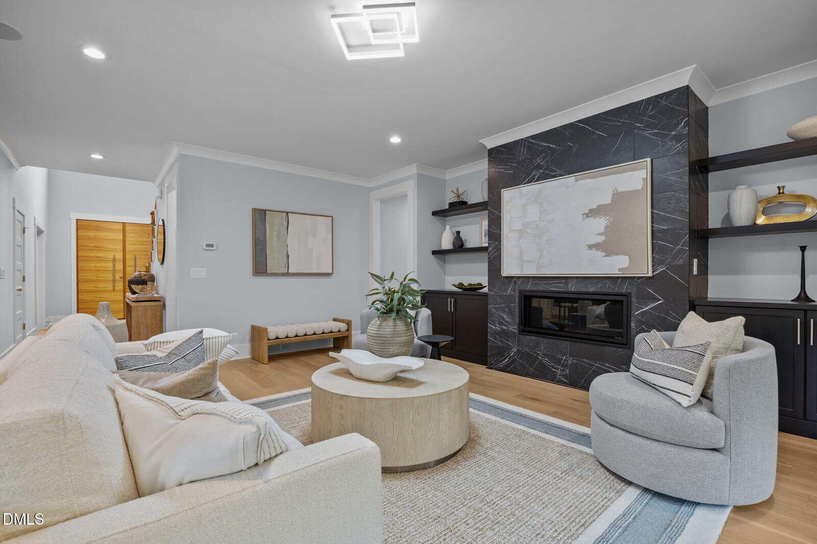 Modern living room with light-colored furniture, round coffee table, black fireplace with marble surround, built-in shelves, decorative objects, large abstract art, and wood flooring. Hallway and entry visible in background.