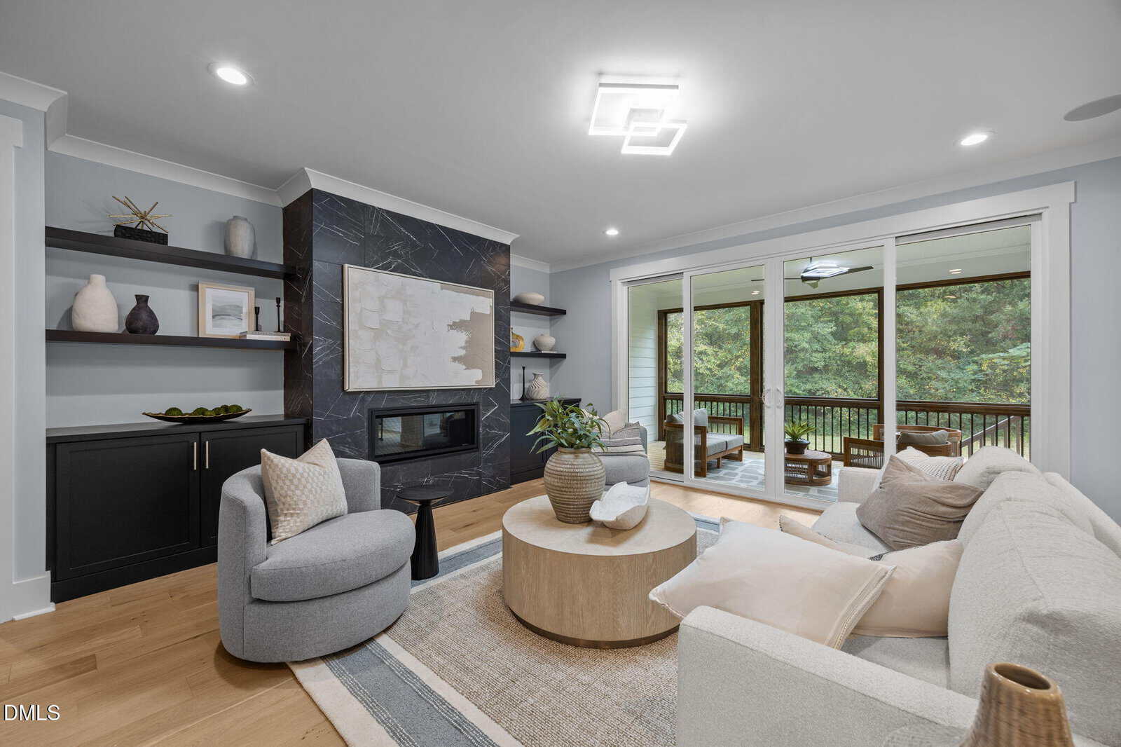 A modern living room with a light gray sofa, round wooden coffee table, armchair, built-in shelves, black fireplace, and large sliding glass doors opening to a screened porch with views of green trees.