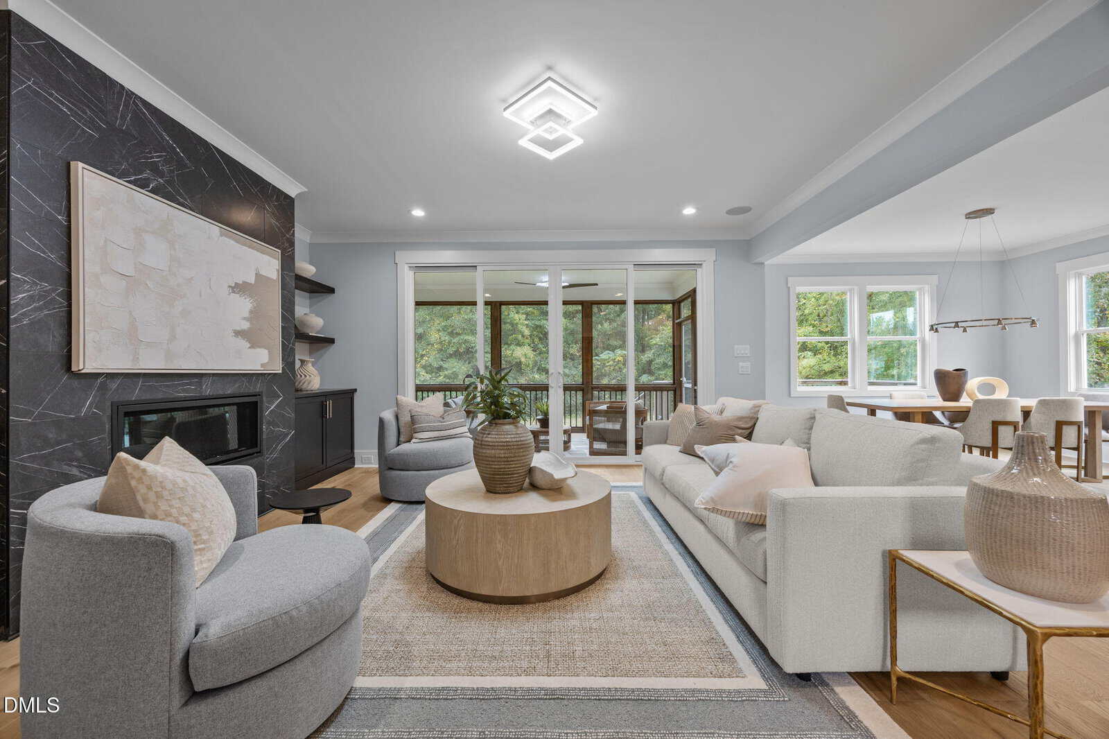 Modern living room with light gray sofas, round wood coffee table, armchair, and large abstract art above a black fireplace. Sliding glass doors open to a screened porch; dining area with wood table is visible on the right.