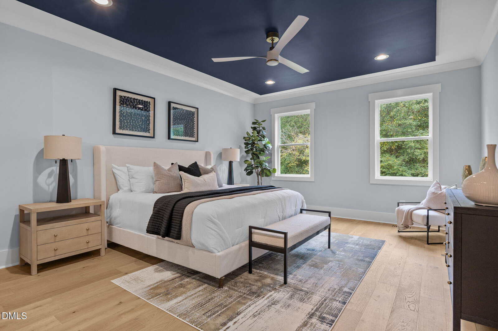 Modern bedroom with a dark blue ceiling, light walls, large bed with neutral bedding, two nightstands with lamps, a bench, area rug, dresser, and chair by two windows overlooking greenery.