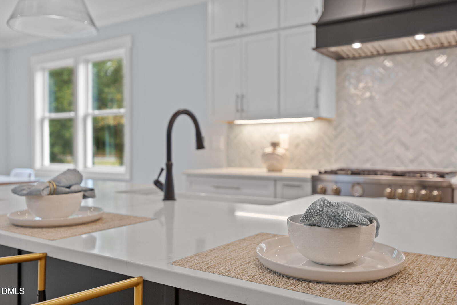 A modern kitchen with a white island countertop, two place settings with white bowls and gray napkins, a black faucet, and a stove with a herringbone tile backsplash in the background.