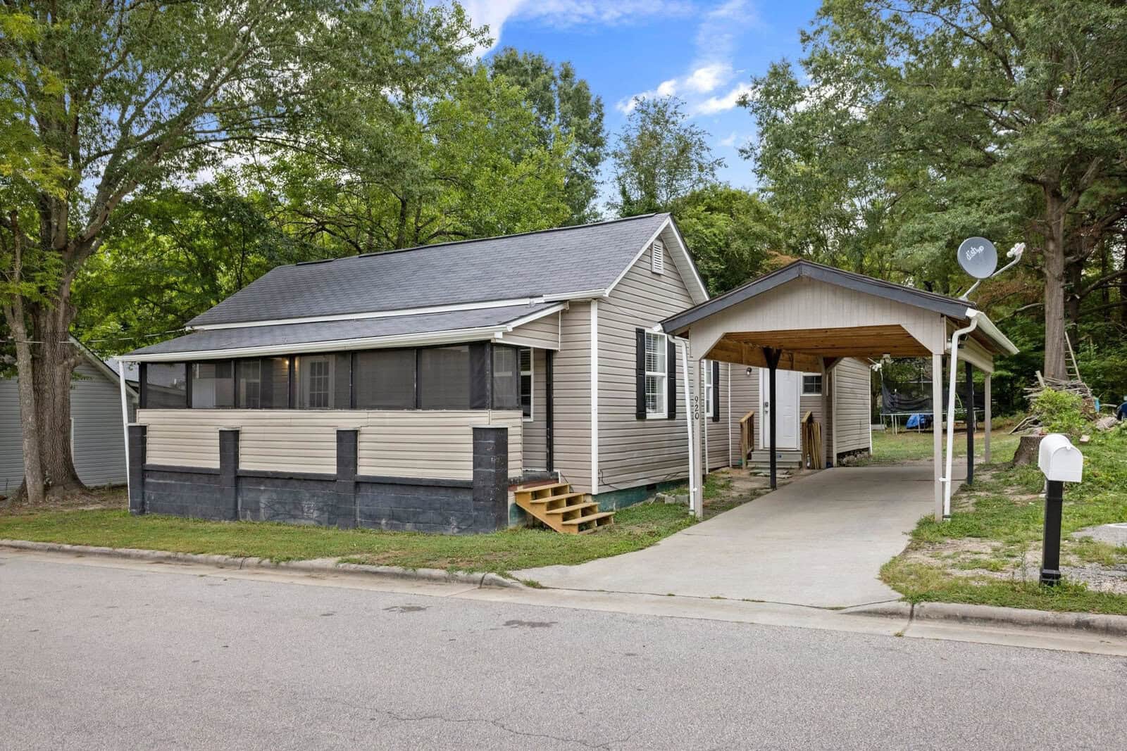 A single-story house at 920 David Street, Henderson NC, with beige siding and a dark trim, featuring a carport on the right and surrounded by trees, with a front yard mailbox by the