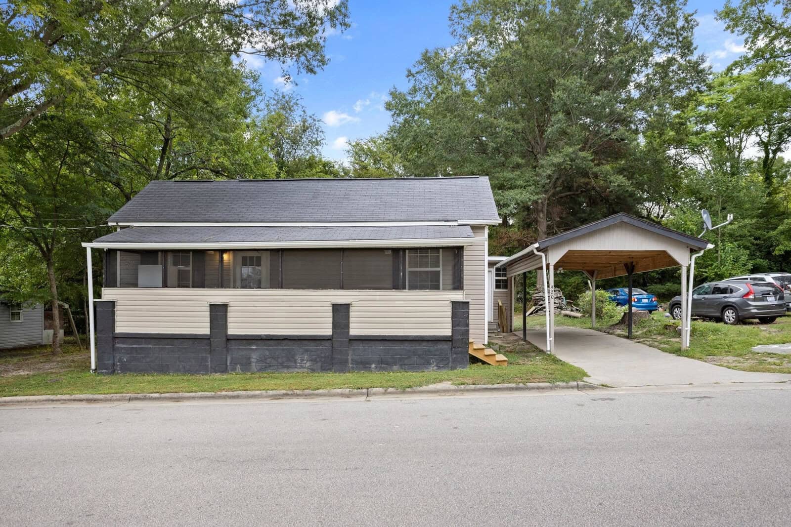 A single-story house with beige siding and a dark gray roof, featuring a carport on the right, is situated beside a street, with trees and a parked car in the background.