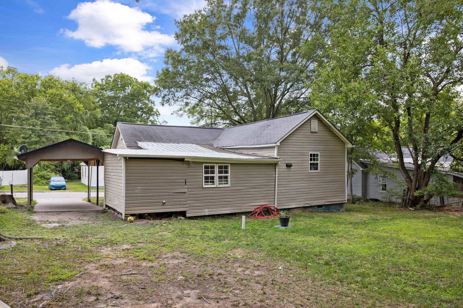 A single-story beige house with gray shingles and white trim, featuring a carport to the left and a sparse lawn with patches of dirt and a single red gardening tool lying on the ground.