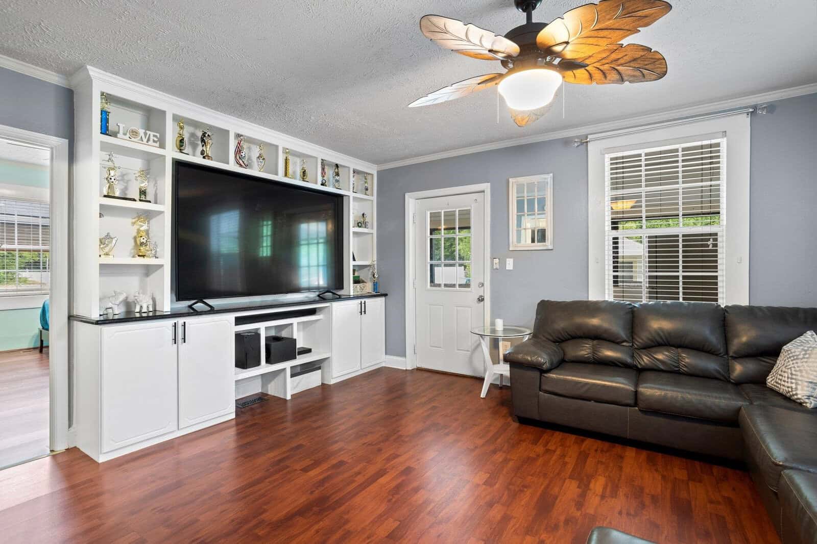 A modern living room with white walls, hardwood flooring, a large wall-mounted television, white cabinetry, and a brown leather couch. a ceiling fan with light wood blades is above, and a doorway leads to another room with a view of a bar-height counter.