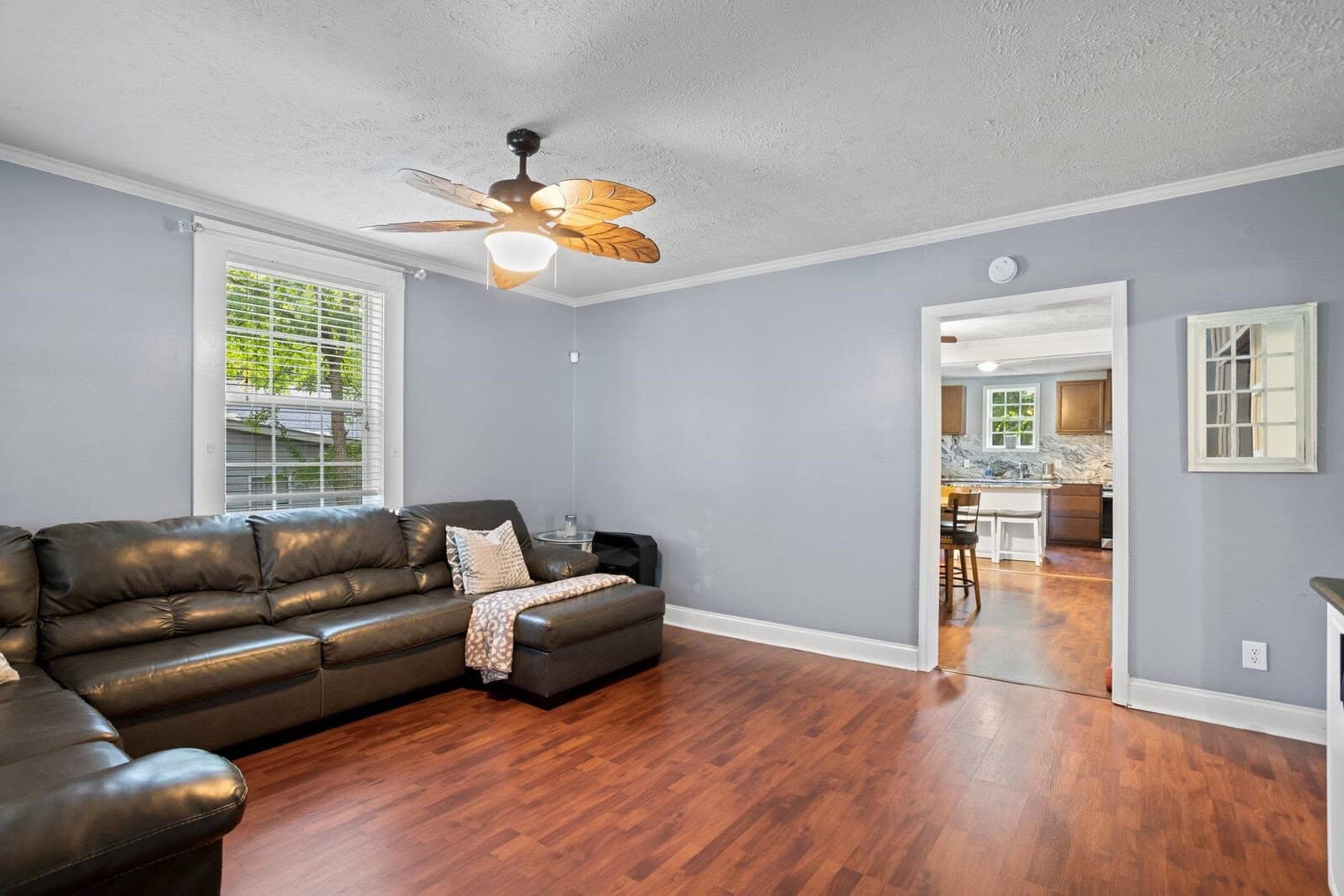 A clean and well-lit living room with a dark brown leather sofa, hardwood floors, and light gray walls, featuring a ceiling fan above and a view into an adjoining room with a kitchen counter.