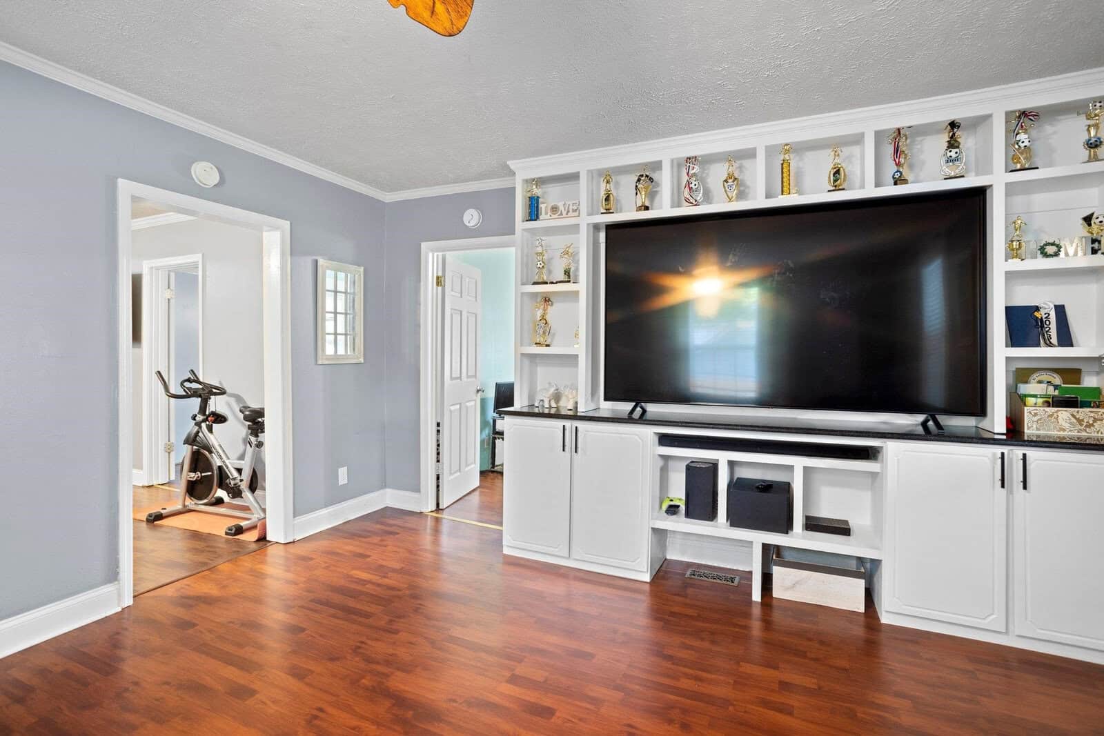 A spacious living room featuring hardwood floors, white cabinetry with a collection of trophies on display, a large flat-screen tv, and an exercise bike in the corner.