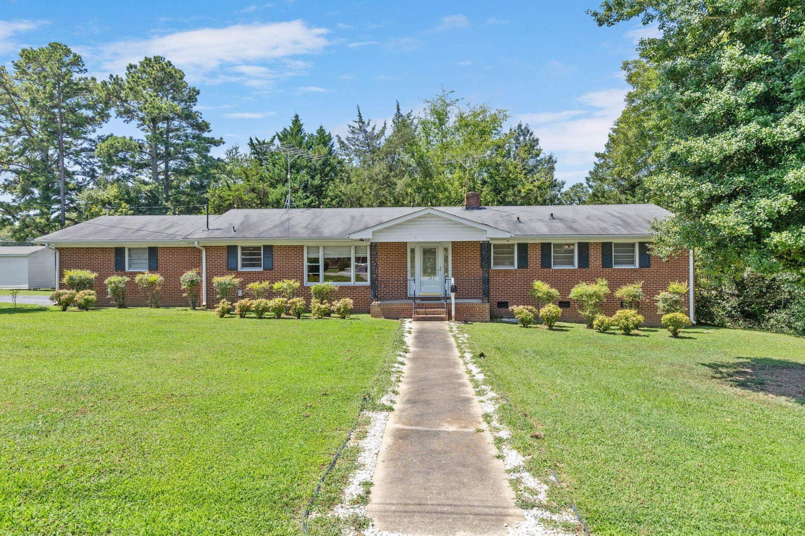 A single-story brick house at 1309 Alpha Drive, Henderson NC, with a shingled roof, featuring white-framed windows and a central white door, is surrounded by a well-maint