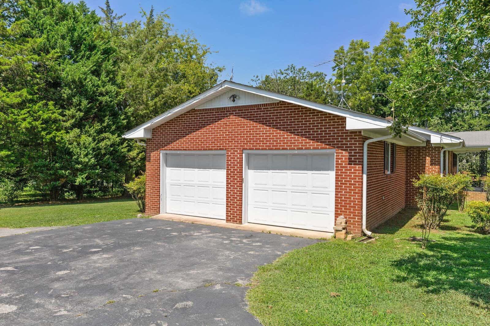 A single-story brick home with a two-car garage and paved driveway, surrounded by green trees under a clear blue sky.
