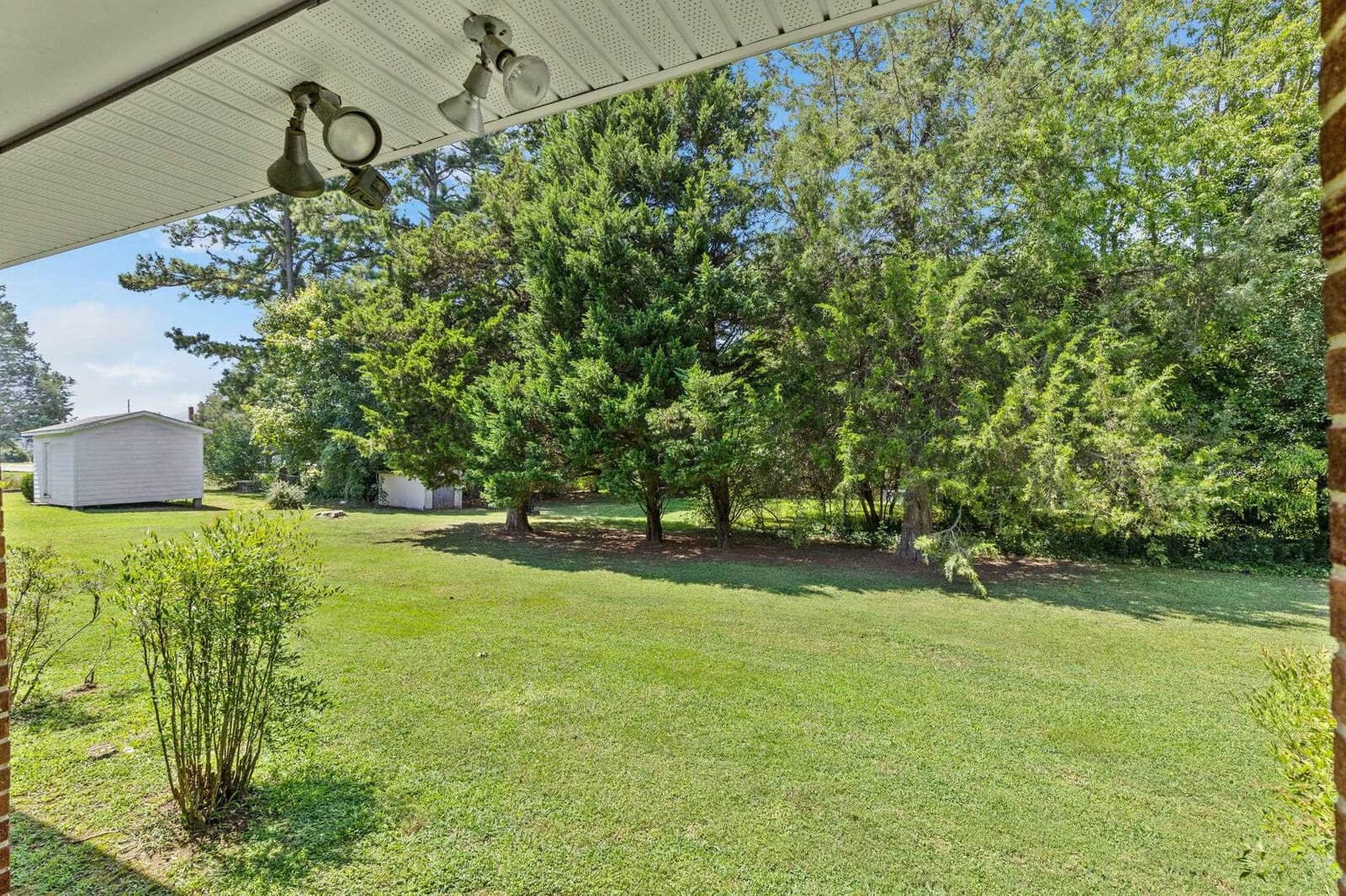 A well-manicured lawn extends towards a cluster of trees with a white shed in the background, as seen from a shaded porch area.