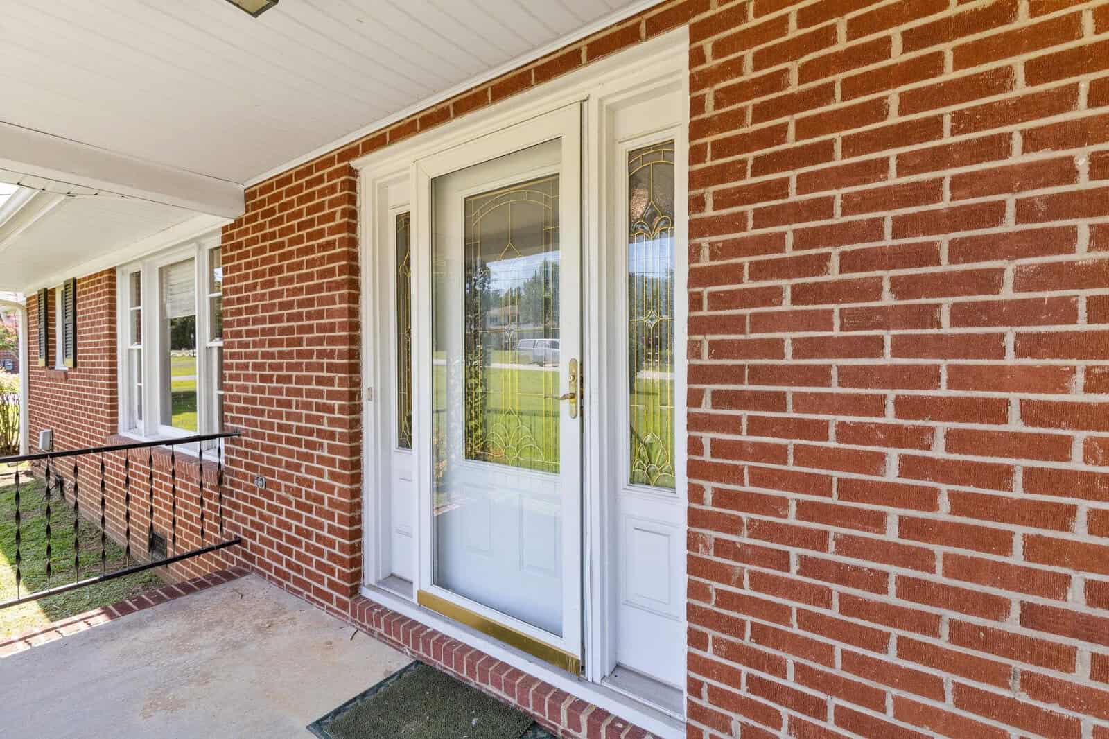 A residential front porch with a white double door featuring glass panels, framed by red brick walls, and a concrete floor, leading out to a sunny yard.