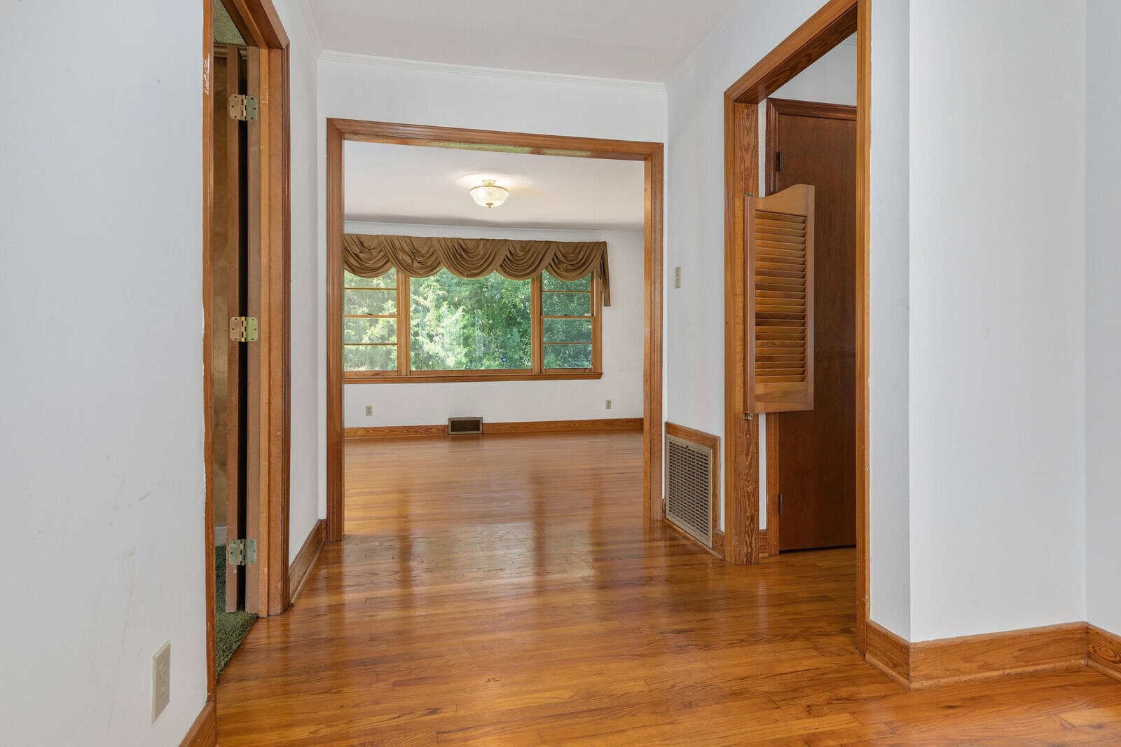 An empty room with hardwood flooring, white walls, and a large window with curtains, viewed from an adjoining doorway.