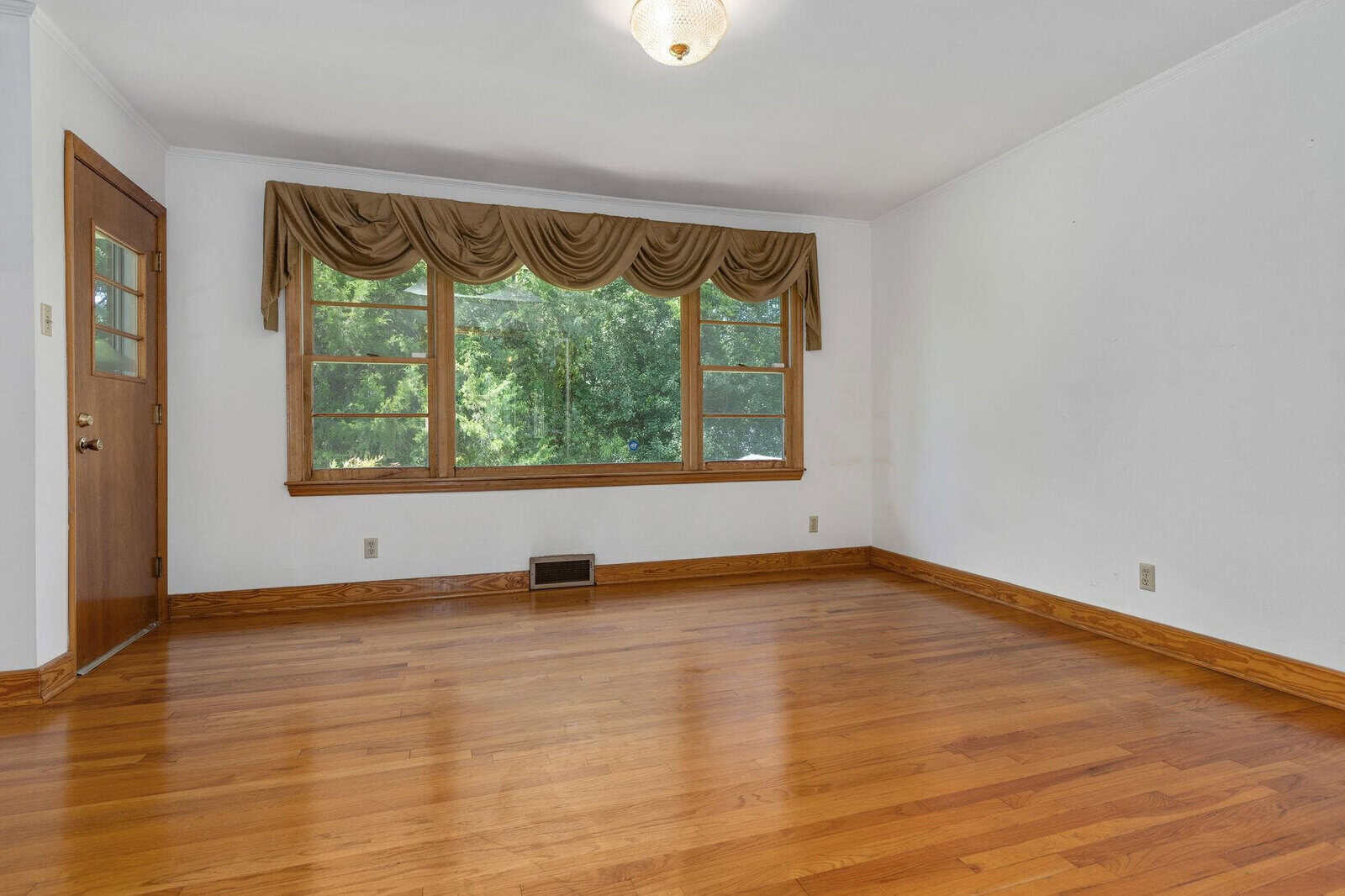 An empty room featuring hardwood floors, a large window with draped valances, and a single door to the side.
