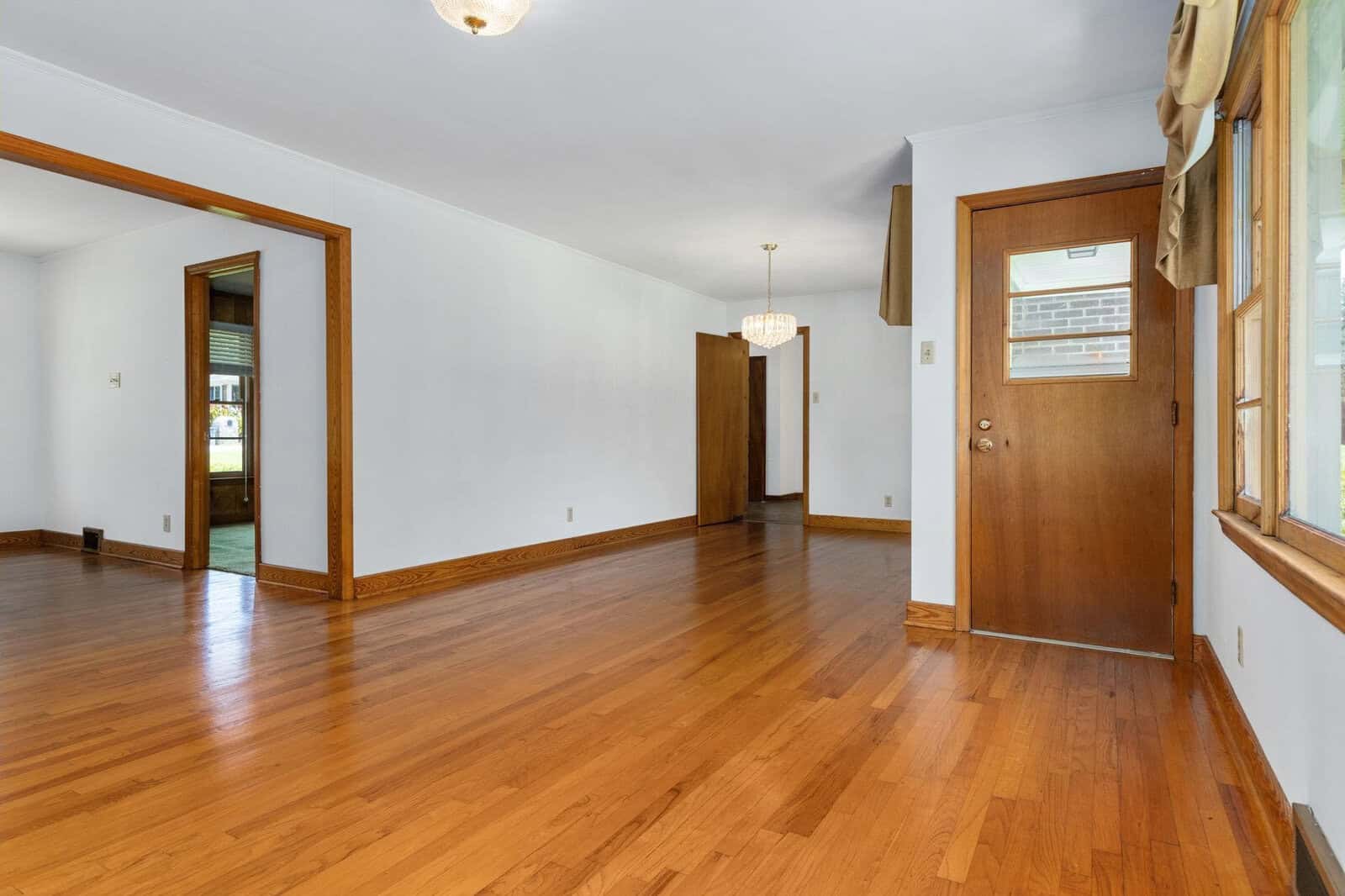 An empty room with polished wooden flooring, white walls, and a window with a partially drawn curtain.