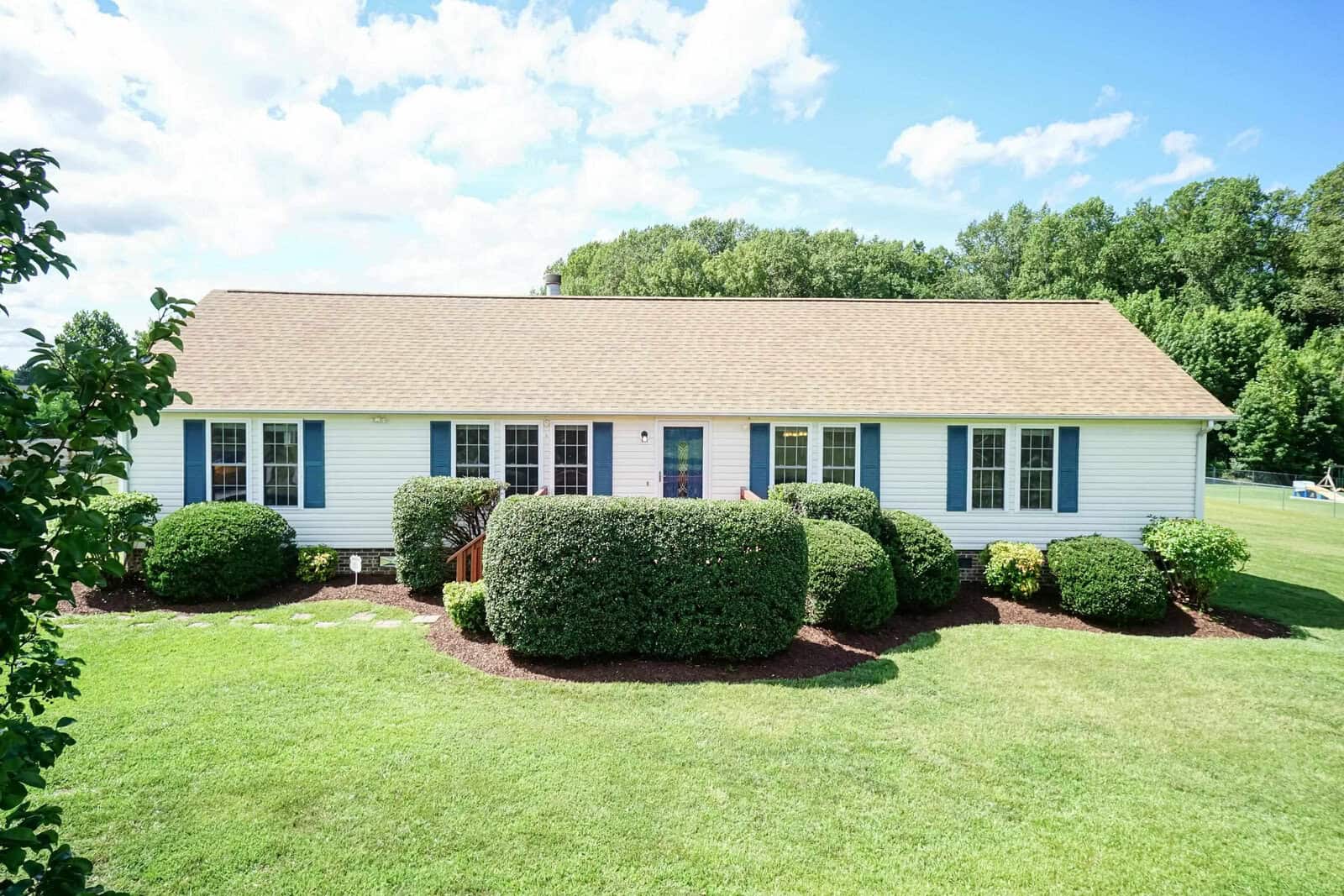 A single-story house with a beige exterior, white trim, and a shingled roof, featuring well-manicured shrubbery in the front yard under a clear sky.
