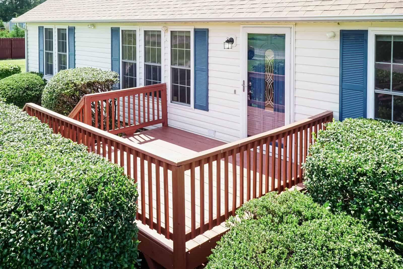 A wooden deck with railing surrounds the entrance to a white house with shutters, nestled within dense shrubbery on a sunny day.