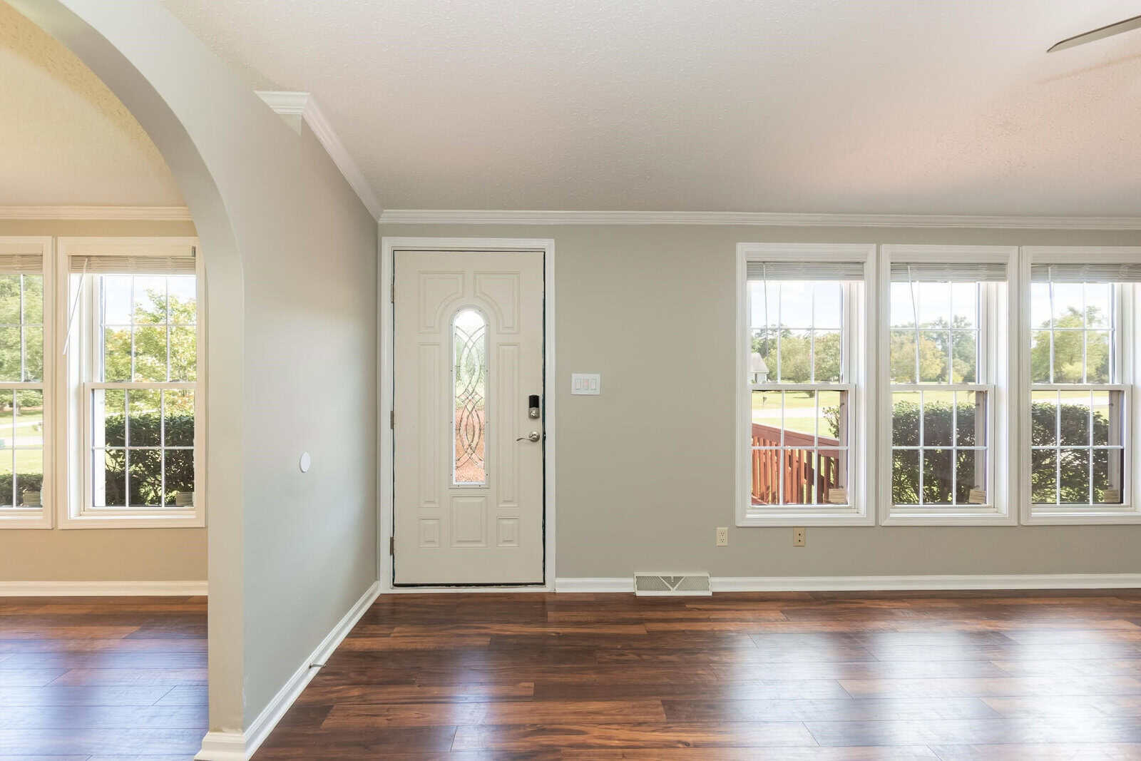 A well-lit, empty room with hardwood flooring, featuring a front door with decorative glass panels and multiple windows overlooking a green landscape.