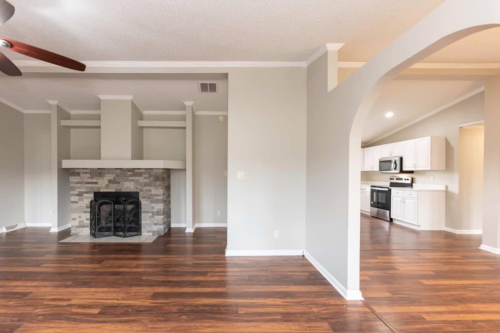 An empty living room with hardwood floors, a fireplace, and an open concept layout leading into a kitchen with modern appliances.