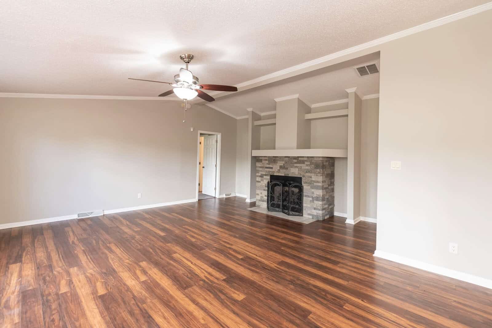An empty living room with polished wooden flooring, a fireplace with a black protective screen, and a ceiling fan illuminated by natural light.