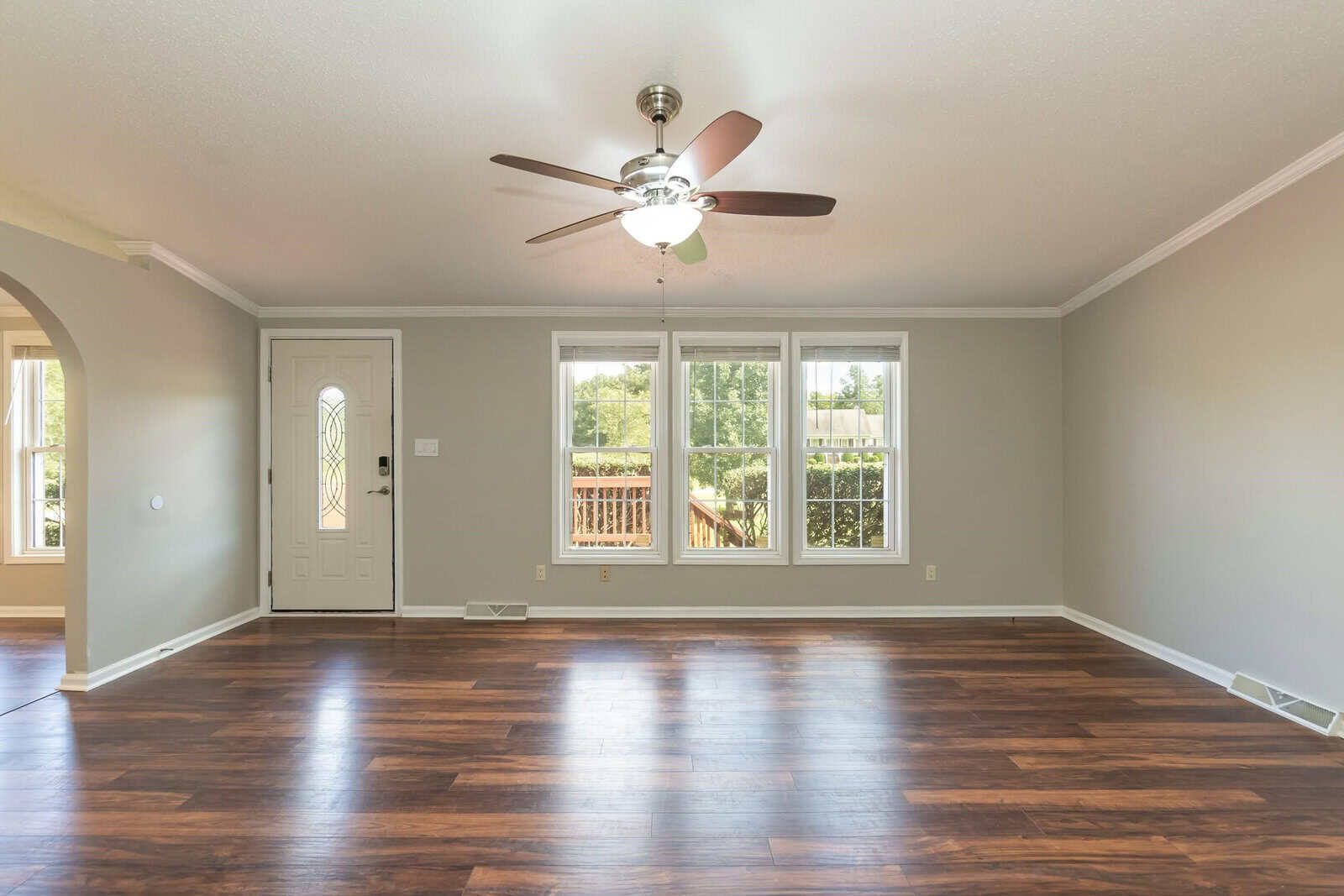 An empty room with polished wooden flooring, a ceiling fan, white walls, and a door leading to a balcony with a view of trees.