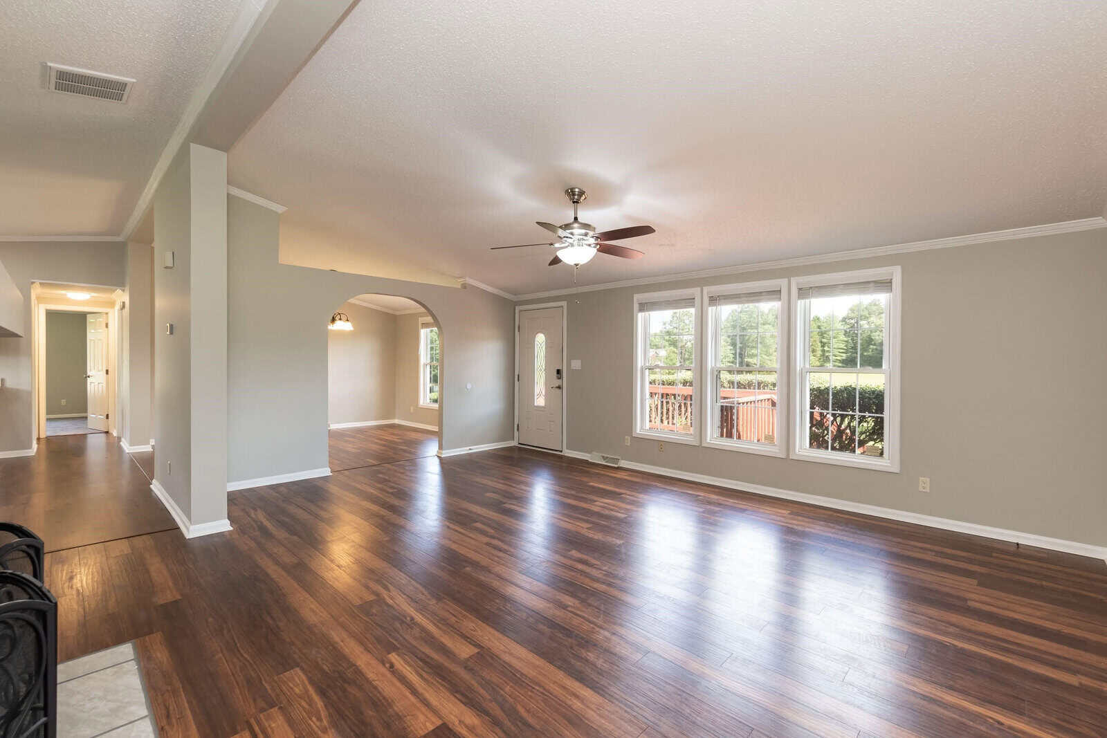 A spacious, empty room with polished hardwood flooring features a ceiling fan, large windows with a view of a deck, and an open doorway leading to another room.